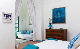 Bright and airy bedroom featuring a white bed with turquoise accents, a wooden dresser, and a cozy seating area in the background. Decorative artwork in blue tones adorns the wall, enhancing the room's serene atmosphere.