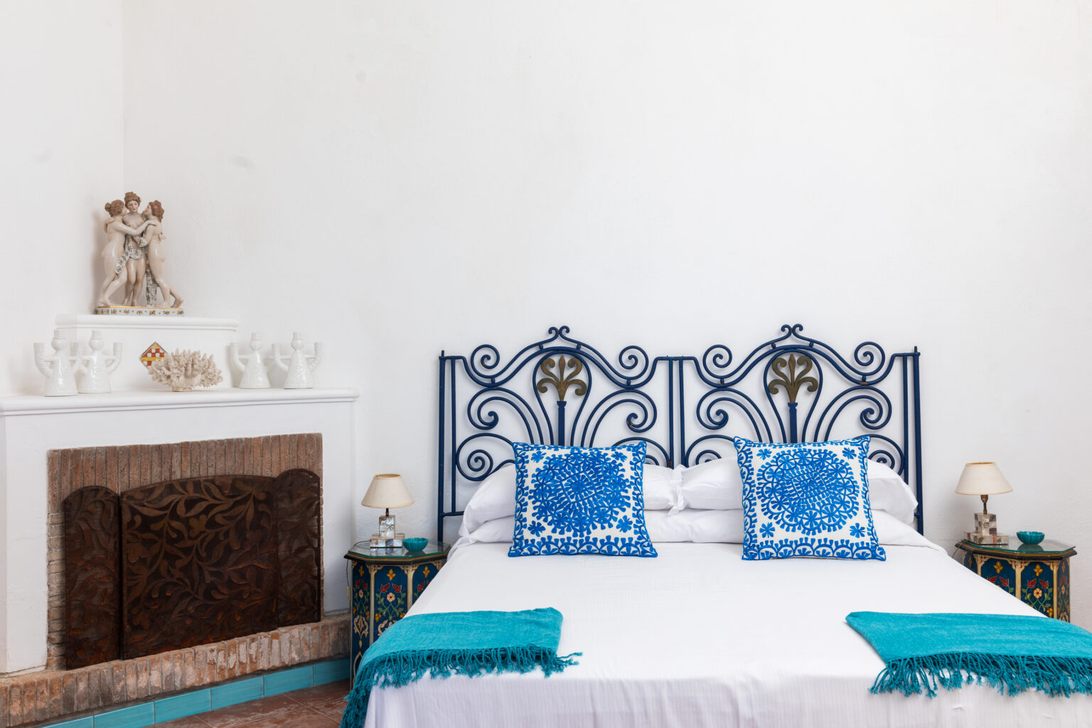 Cozy bedroom featuring a wrought iron bed frame with blue decorative pillows, a white wall, and a stylish fireplace. The room exudes a serene and inviting atmosphere, ideal for relaxation. | Only on SopranoVillas