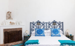 Cozy bedroom featuring a wrought iron bed frame with blue decorative pillows, a white wall, and a stylish fireplace. The room exudes a serene and inviting atmosphere, ideal for relaxation.