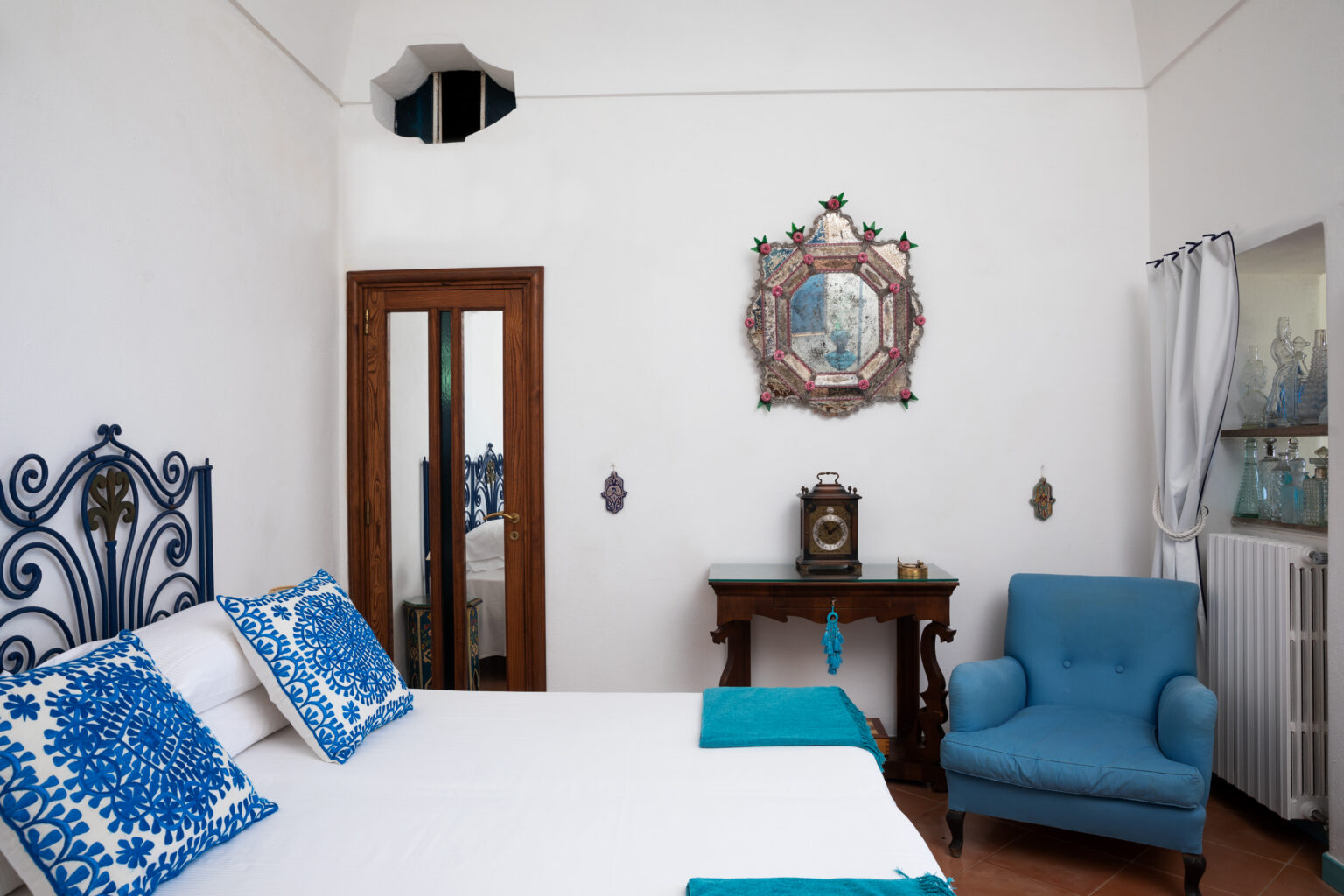 Cozy bedroom featuring a white bed with blue decorative pillows, a wooden dresser, a vintage clock, and a blue armchair, set against a bright white wall with artistic decor. | Only on SopranoVillas