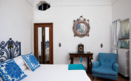 Cozy bedroom featuring a white bed with blue decorative pillows, a wooden dresser, a vintage clock, and a blue armchair, set against a bright white wall with artistic decor.