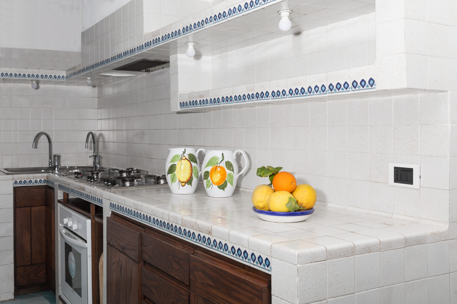 Modern kitchen with white tiled walls and wooden cabinets, featuring a gas stove, sink, and decorative pitchers with lemons and oranges on the countertop. | Only on SopranoVillas
