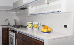Modern kitchen with white tiled walls and wooden cabinets, featuring a gas stove, sink, and decorative pitchers with lemons and oranges on the countertop.