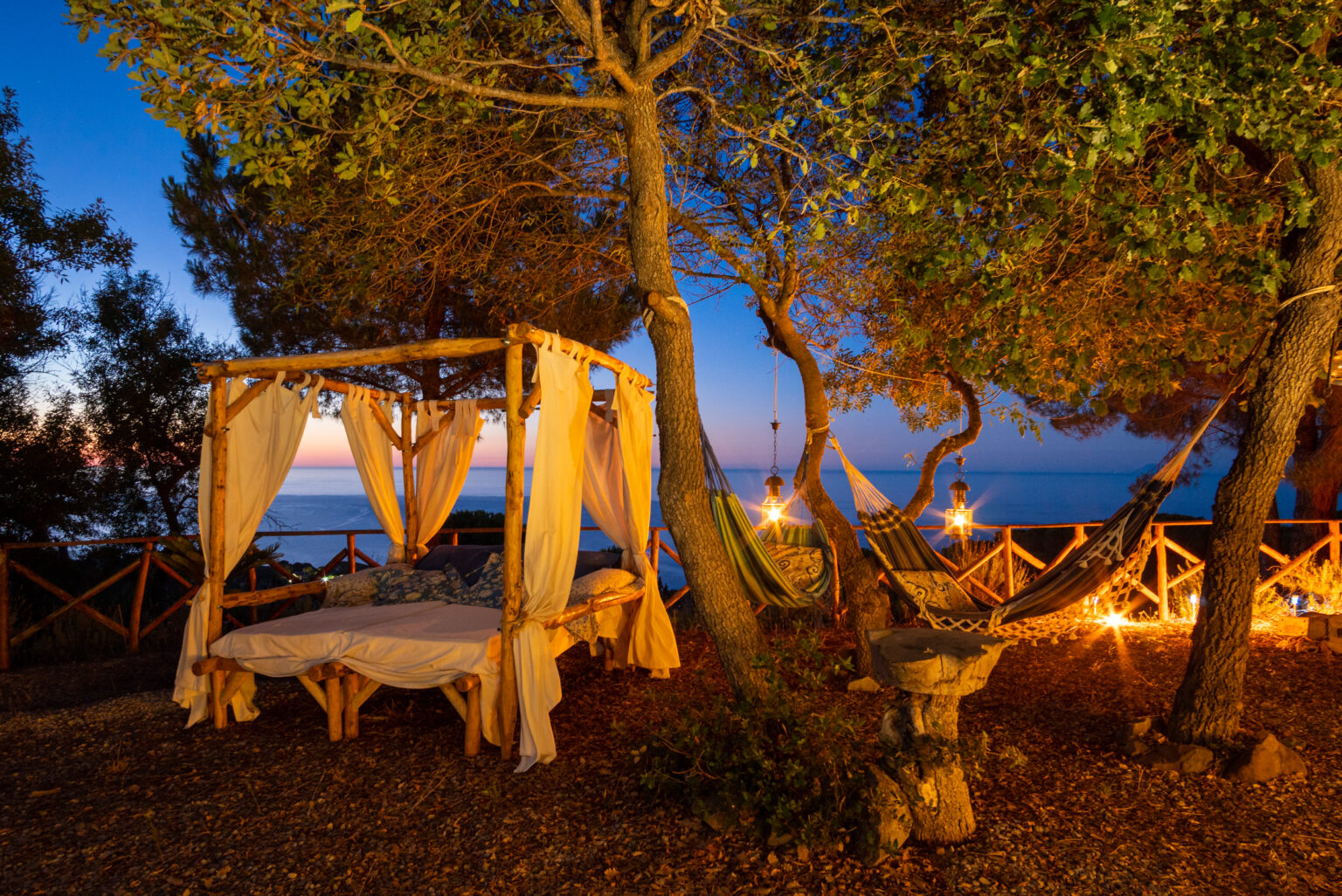 Romantic outdoor setting with a canopy bed and hammocks under trees at sunset, overlooking the ocean, creating a serene and inviting atmosphere. | Only on SopranoVillas