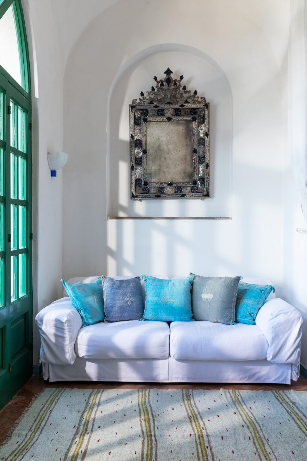 Cozy living room with a white sofa adorned with blue and gray decorative pillows, complemented by a vintage wall mirror and green window, creating a bright and inviting atmosphere. | Only on SopranoVillas