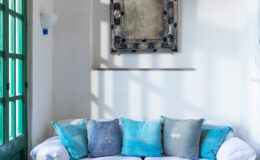 Cozy living room with a white sofa adorned with blue and gray decorative pillows, complemented by a vintage wall mirror and green window, creating a bright and inviting atmosphere.