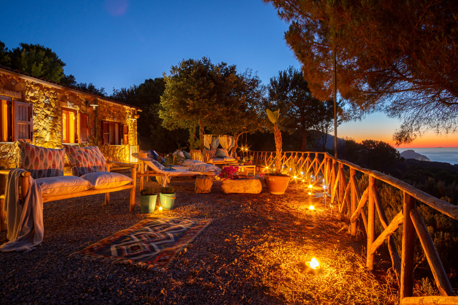 Cozy outdoor seating area with wooden furniture and decorative pillows, illuminated by candles at sunset, overlooking a scenic landscape with trees and a warm sky. Perfect for relaxation and enjoying nature. | Only on SopranoVillas