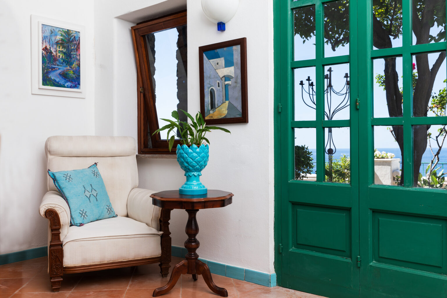 Cozy living space featuring a comfortable cream armchair with a turquoise decorative pillow, a wooden side table, and a vibrant turquoise planter with green foliage. The room is illuminated by natural light from a large green-framed door, leading to a scenic view of the outdoors and a painted artwork on the wall. | Only on SopranoVillas