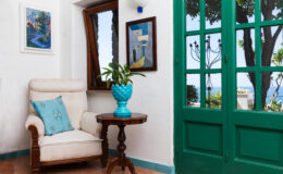 Cozy living space featuring a comfortable cream armchair with a turquoise decorative pillow, a wooden side table, and a vibrant turquoise planter with green foliage. The room is illuminated by natural light from a large green-framed door, leading to a scenic view of the outdoors and a painted artwork on the wall.