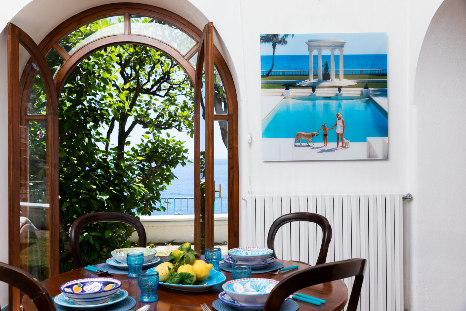 Bright dining area featuring a round wooden table set with colorful plates and fresh fruit, framed by an arched window showcasing a lush garden. A vibrant painting of a pool scene hangs on the wall, adding a touch of elegance to the space. | Only on SopranoVillas