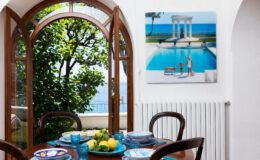 Bright dining area featuring a round wooden table set with colorful plates and fresh fruit, framed by an arched window showcasing a lush garden. A vibrant painting of a pool scene hangs on the wall, adding a touch of elegance to the space.