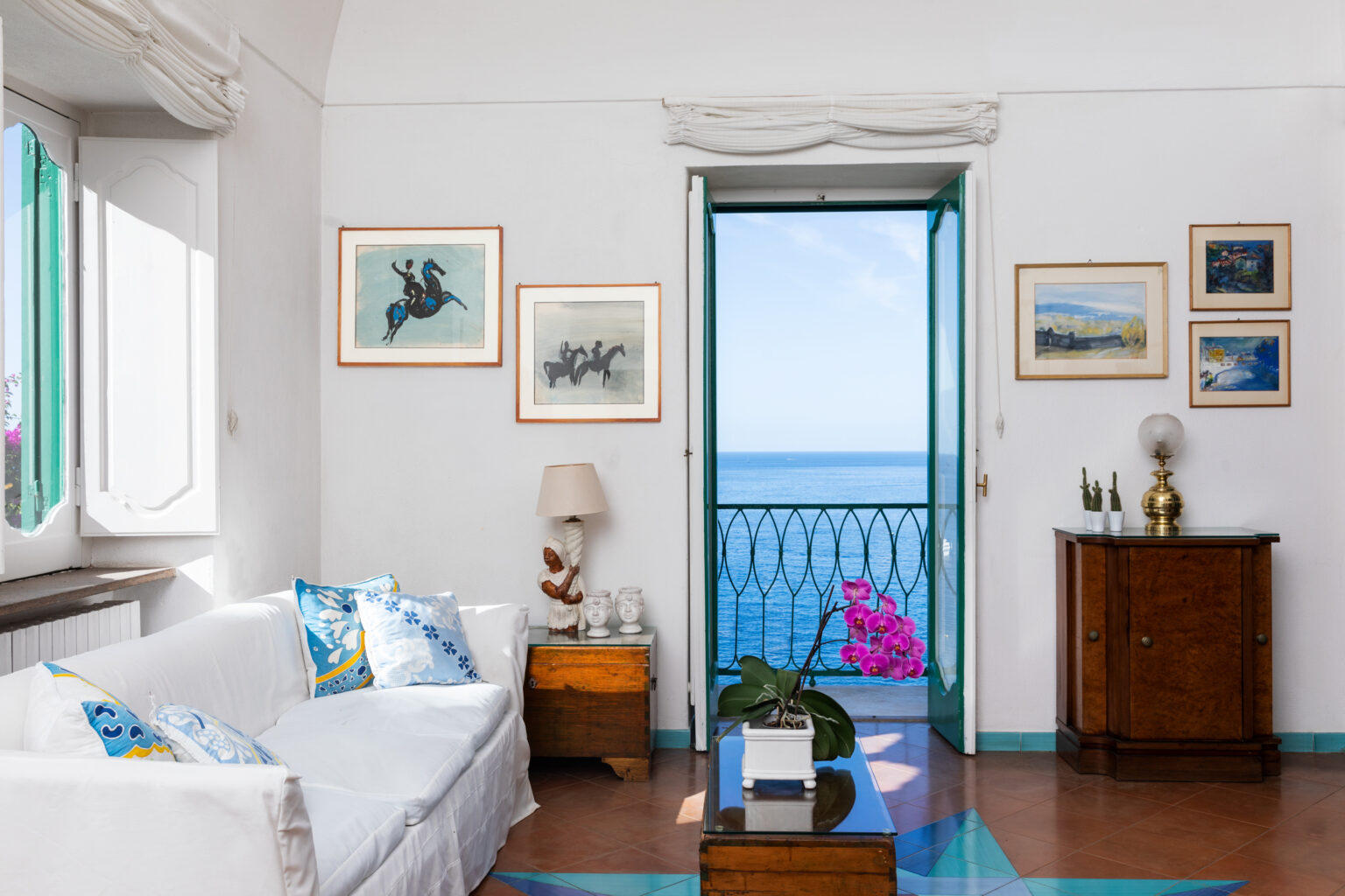 Bright and airy living room with a white sofa and colorful cushions, featuring artwork on the walls and a view of the sea through an open door. A potted plant and wooden furniture add warmth to the space, creating a serene coastal atmosphere. | Only on SopranoVillas