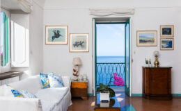 Bright and airy living room with a white sofa and colorful cushions, featuring artwork on the walls and a view of the sea through an open door. A potted plant and wooden furniture add warmth to the space, creating a serene coastal atmosphere.