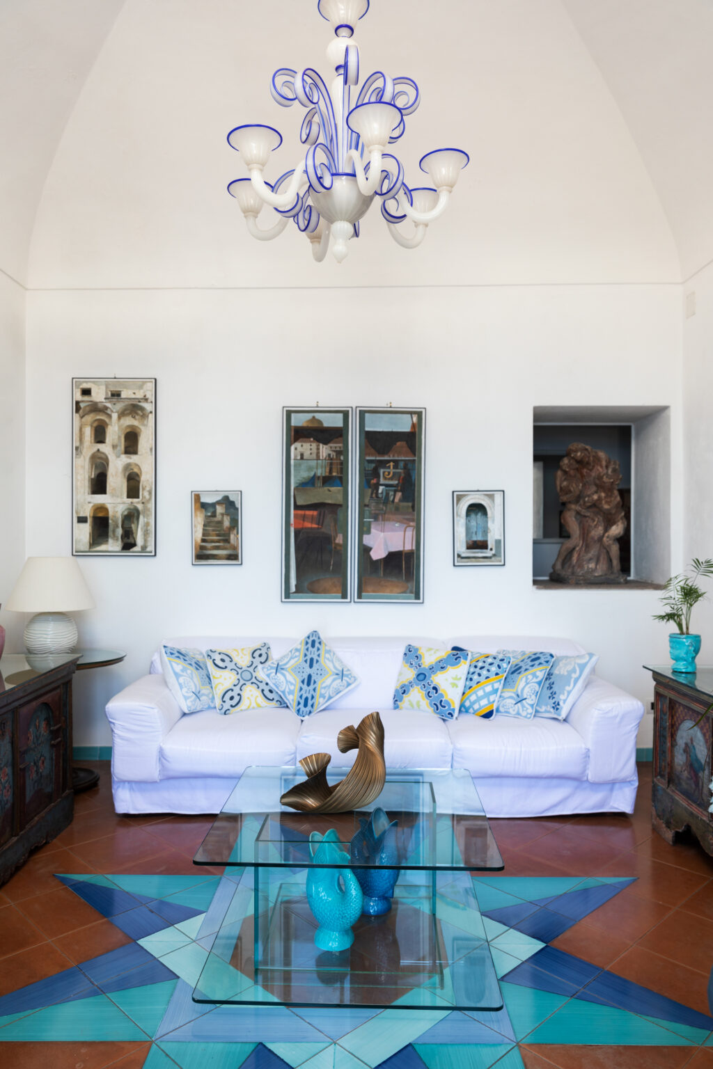 Bright and stylish living room featuring a white sofa adorned with decorative pillows, a glass coffee table with artistic sculptures, and a colorful tiled floor design. The room is illuminated by a unique chandelier and showcases various artworks on the walls, creating an inviting and modern atmosphere. | Only on SopranoVillas