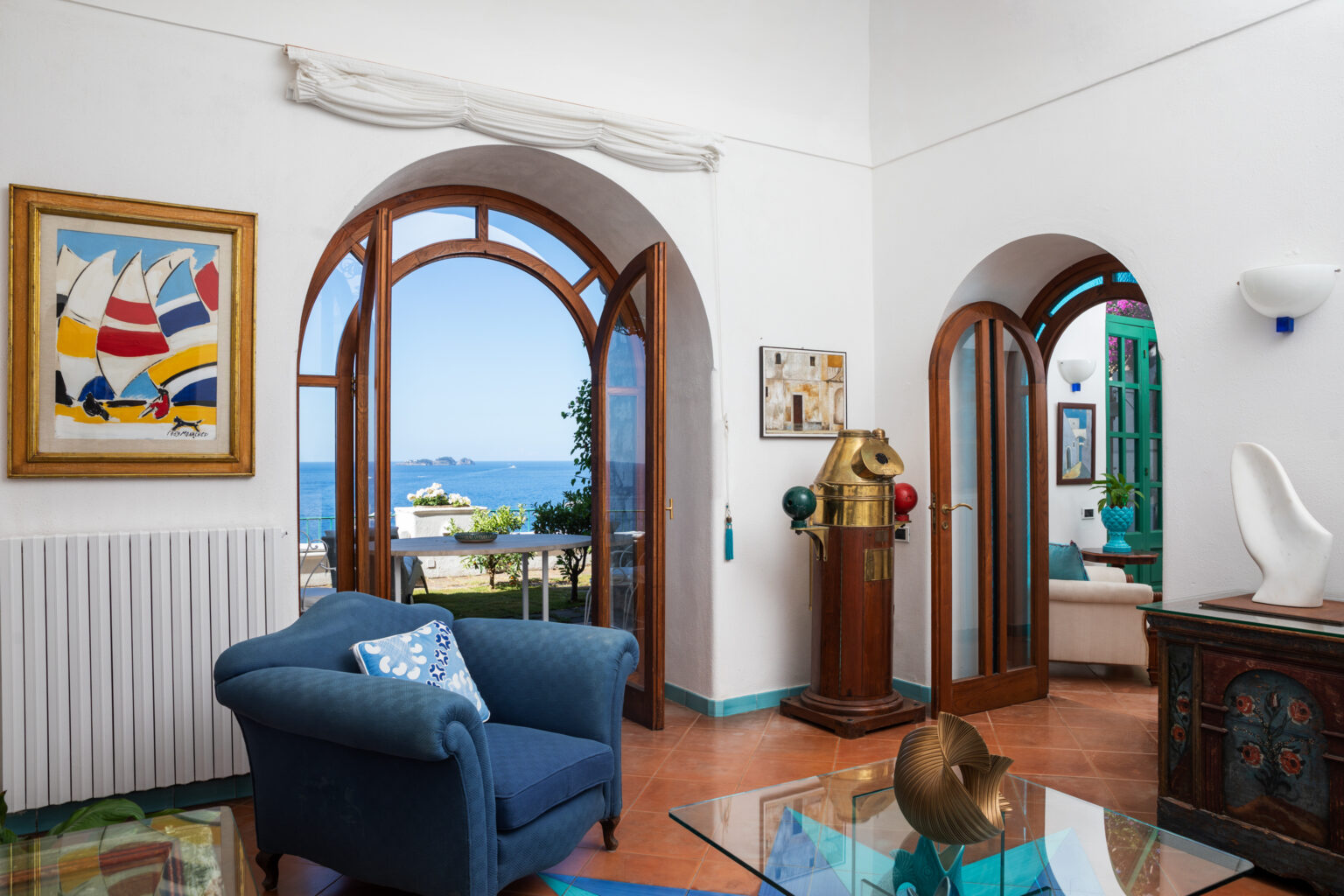 Bright and airy coastal living room featuring arched wooden windows, vibrant artwork, and a scenic view of the sea. The space is decorated with a blue sofa, a decorative side table, and unique furnishings, creating a welcoming atmosphere. | Only on SopranoVillas
