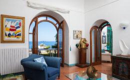 Bright and airy coastal living room featuring arched wooden windows, vibrant artwork, and a scenic view of the sea. The space is decorated with a blue sofa, a decorative side table, and unique furnishings, creating a welcoming atmosphere.