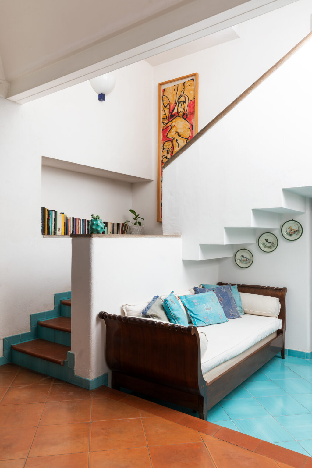 Cozy interior of a modern home featuring a wooden sofa adorned with colorful cushions, a bookshelf with books, decorative plates on the wall, and a staircase leading to the upper level. The walls are painted white, enhancing the bright and airy ambiance. | Only on SopranoVillas