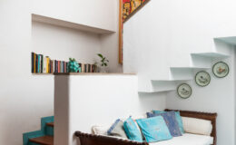 Cozy interior of a modern home featuring a wooden sofa adorned with colorful cushions, a bookshelf with books, decorative plates on the wall, and a staircase leading to the upper level. The walls are painted white, enhancing the bright and airy ambiance.