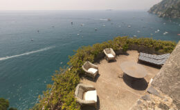 Scenic view of a coastal terrace overlooking the turquoise sea, featuring comfortable wicker chairs and a round table, with boats sailing in the distance and rocky cliffs in the background. Ideal for relaxation and enjoying breathtaking ocean views.