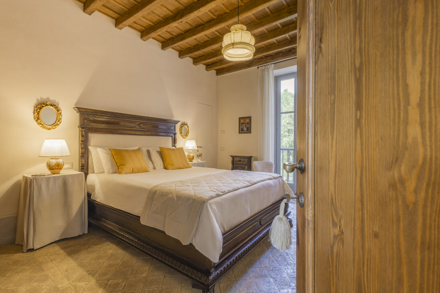 Cozy bedroom interior featuring a large bed with white linens and yellow accent pillows, wooden ceiling beams, bedside lamps, and a view of a window with natural light. | Only on SopranoVillas
