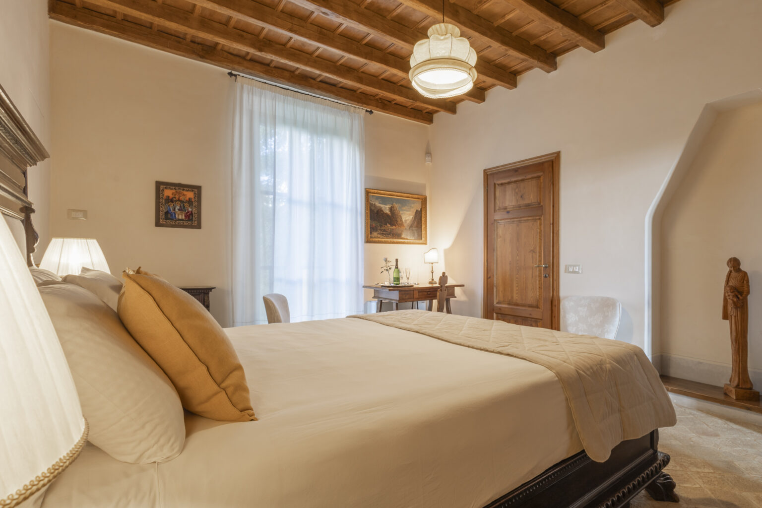 Cozy bedroom featuring a wooden ceiling, a large window with sheer curtains, and a comfortable bed adorned with decorative pillows. The room includes a wooden door, a small table with a bottle and glasses, and a decorative statue, creating a warm and inviting atmosphere. Ideal for relaxation and comfort. | Only on SopranoVillas
