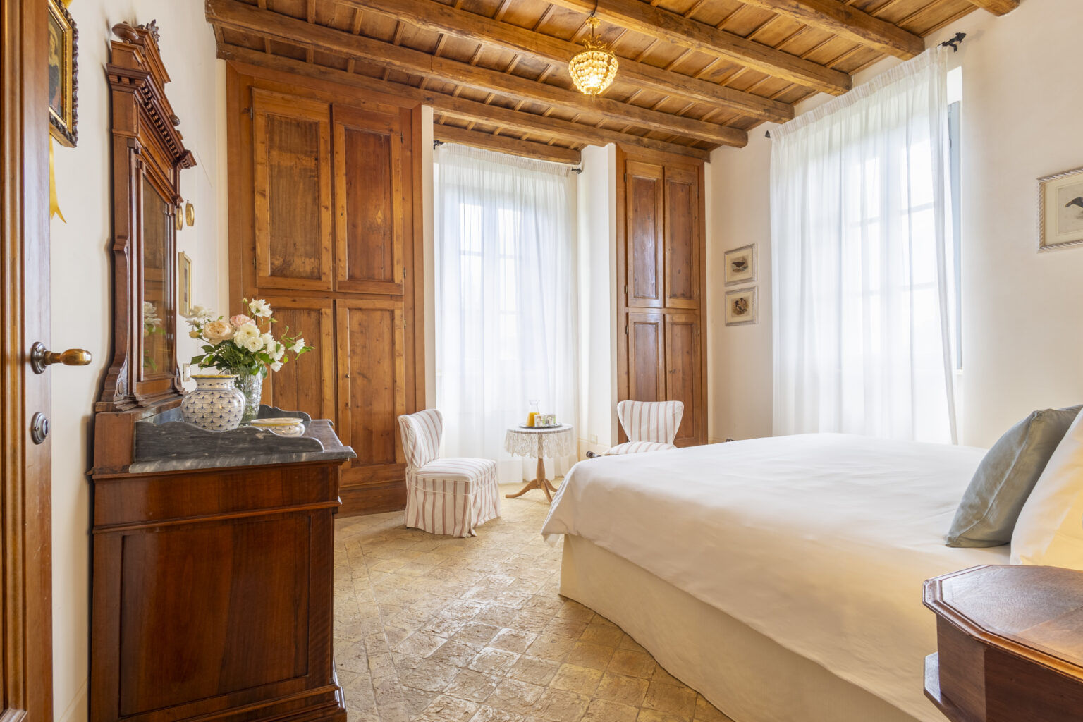 Cozy bedroom with wooden ceiling beams, large windows allowing natural light, and elegant furnishings including a bed, chairs, and a vintage dresser, creating a warm and inviting atmosphere. | Only on SopranoVillas