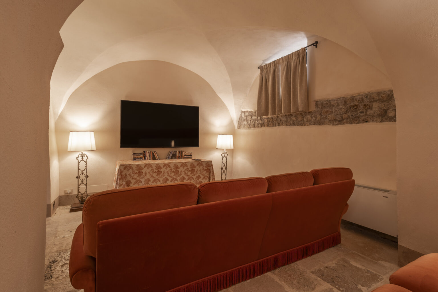 Cozy living room with an arched ceiling, featuring a large flat-screen TV, two stylish lamps, and a comfortable orange sofa, creating a warm and inviting atmosphere. | Only on SopranoVillas