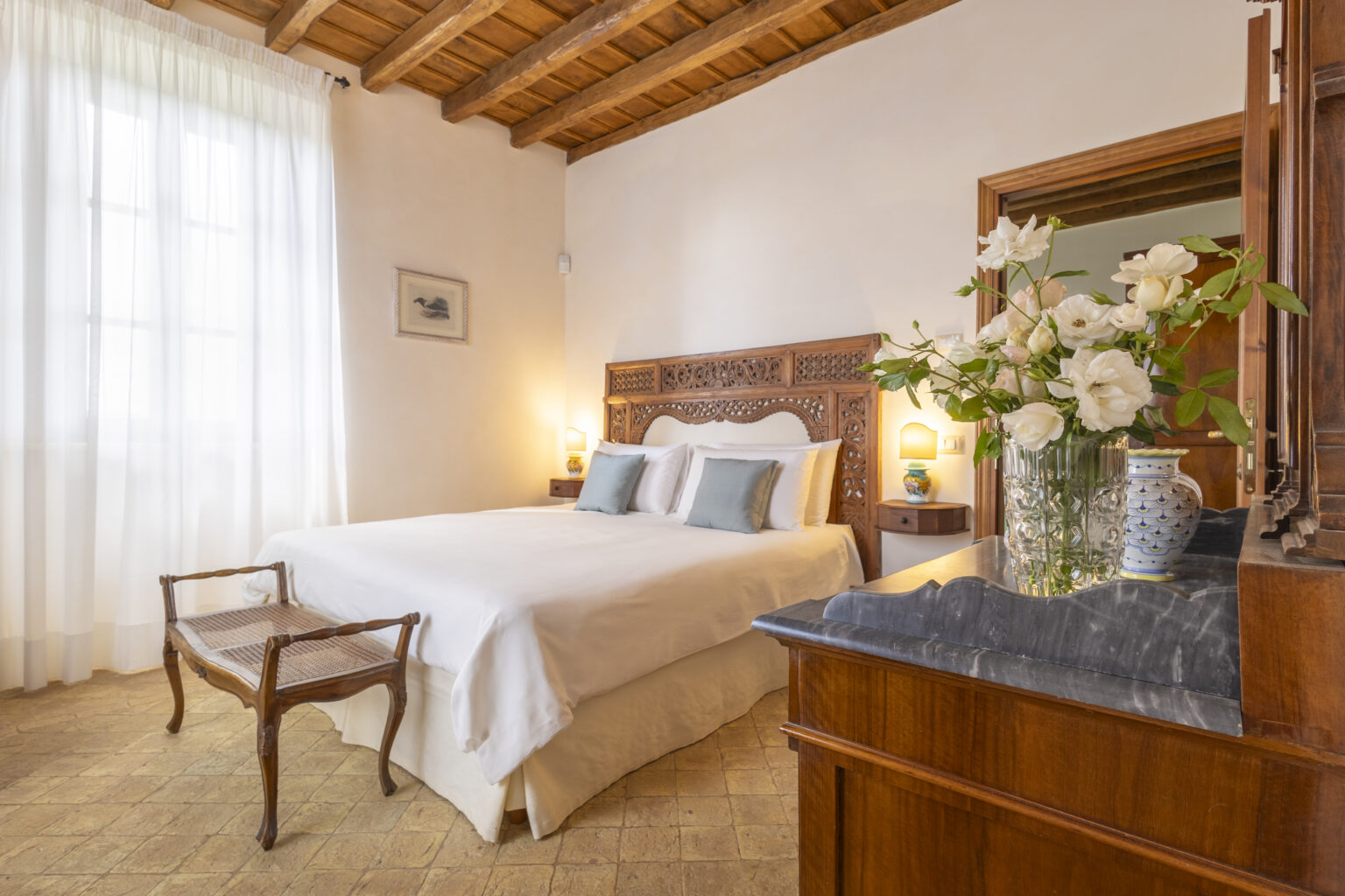 Cozy bedroom featuring a wooden bed with white linens, elegant decor, a bedside table with a vase of flowers, and large windows allowing natural light to brighten the space. | Only on SopranoVillas