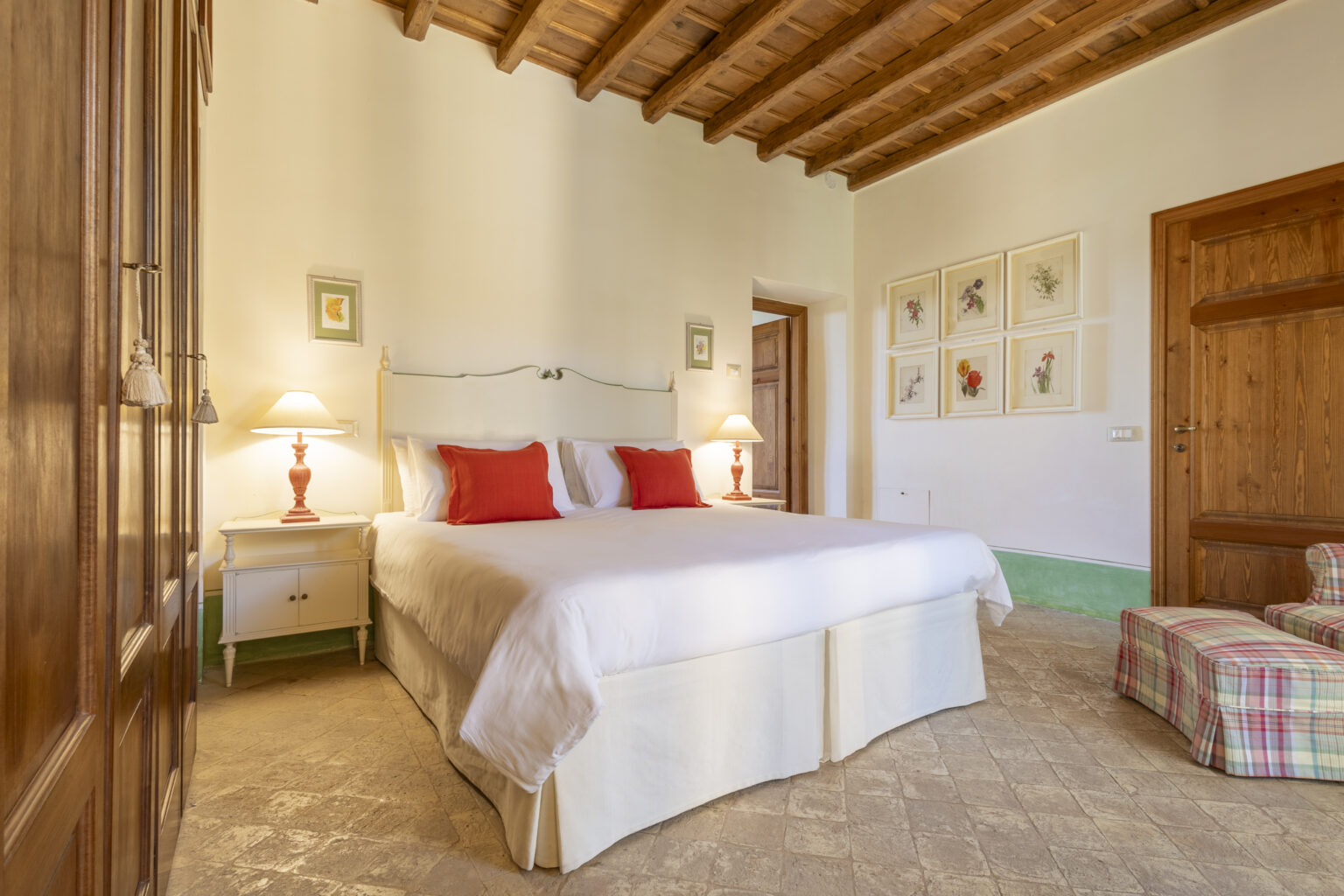 Cozy bedroom featuring a queen-sized bed with white linens and red accent pillows, wooden beams on the ceiling, and soft lighting from bedside lamps. The room includes a wooden door and a small seating area with a plaid ottoman, complemented by framed botanical artwork on the walls. | Only on SopranoVillas