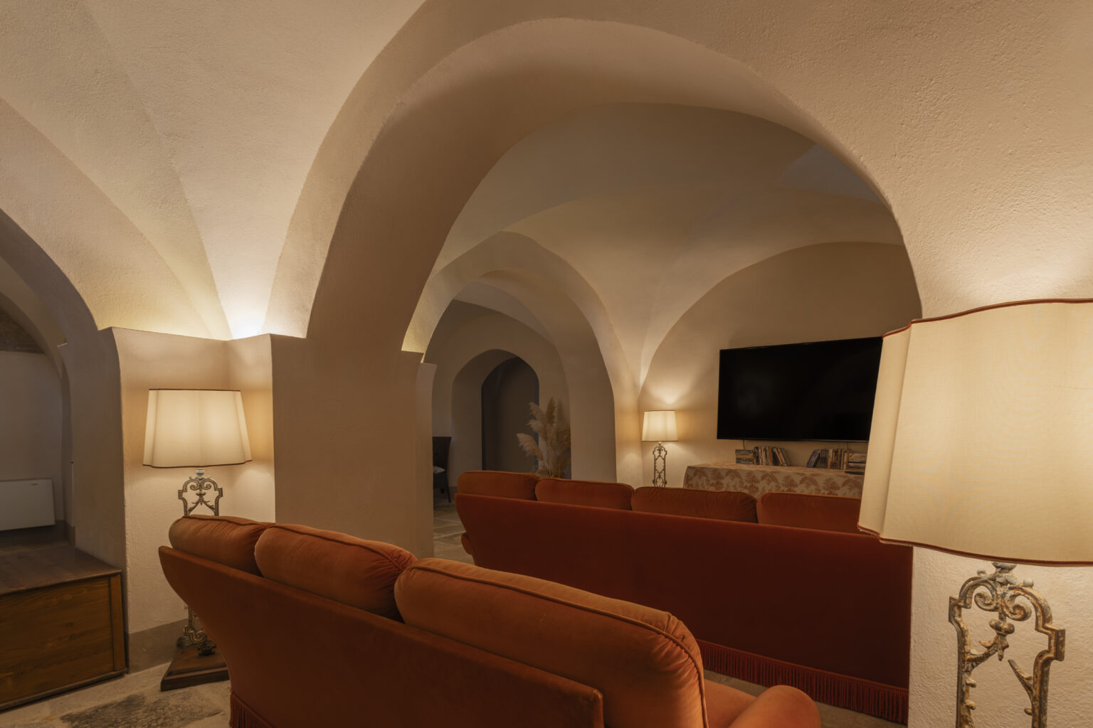 Cozy living room with arched ceilings, soft lighting, and plush orange sofas, featuring a modern TV and decorative lamps that enhance the warm ambiance. | Only on SopranoVillas