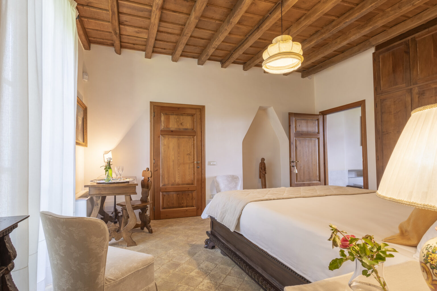 Cozy bedroom featuring wooden ceiling beams, a large bed with neutral bedding, a bedside lamp, and a small table with a vase of flowers, creating a warm and inviting atmosphere. | Only on SopranoVillas