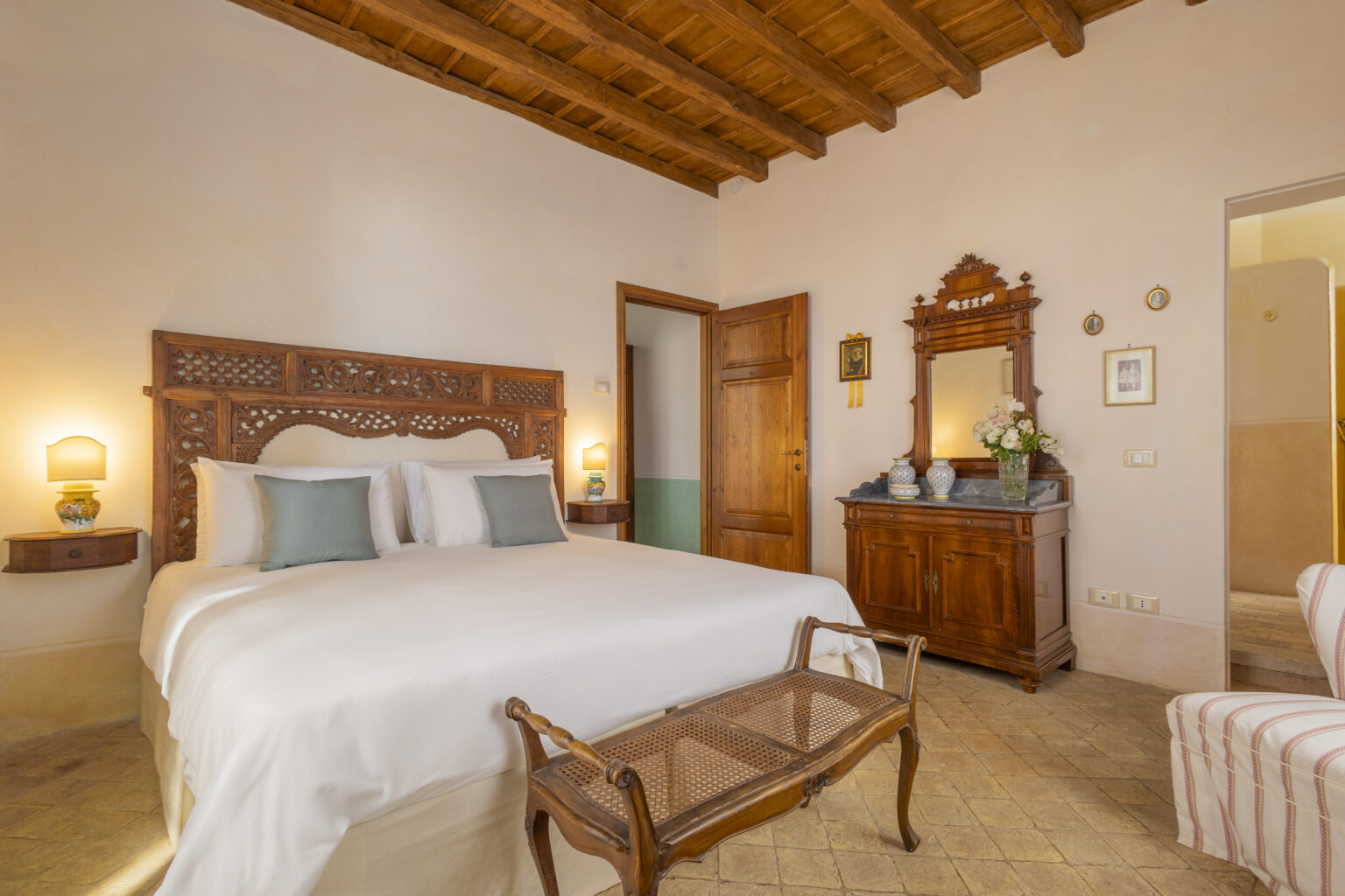 Cozy bedroom featuring a queen-sized bed with decorative headboard, a wooden dresser, and a vintage mirror, illuminated by soft lighting and adorned with warm, earthy tones. | Only on SopranoVillas