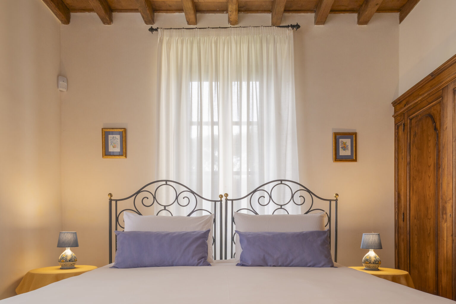 Cozy bedroom featuring a wrought iron bed with purple pillows, soft lighting from bedside lamps, and elegant wooden furniture, complemented by sheer curtains and framed artwork. Perfect for a relaxing retreat. | Only on SopranoVillas