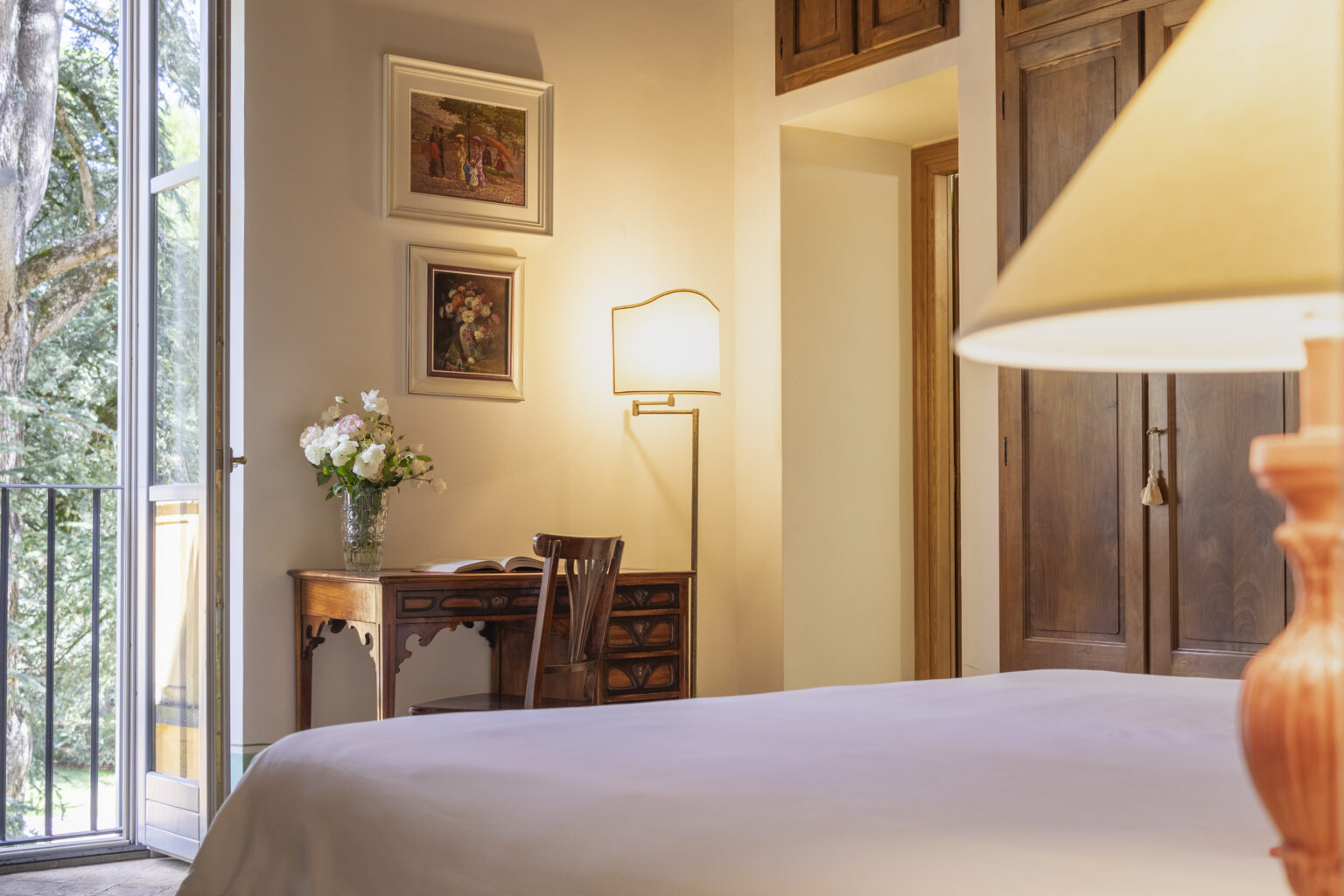 Cozy bedroom interior featuring a comfortable bed, a wooden desk with a vase of flowers, and warm lighting from a bedside lamp, with a view of a balcony and framed artwork on the walls. Ideal for relaxation and work. | Only on SopranoVillas