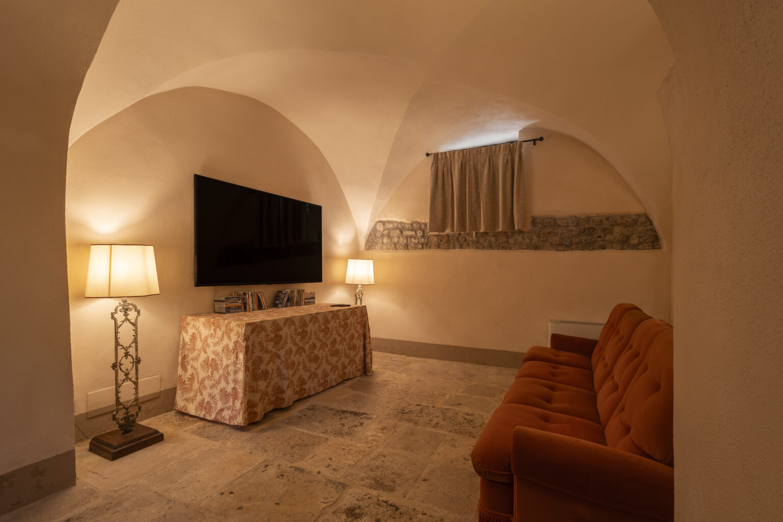 Cozy living room with arched ceilings, featuring an orange velvet sofa, a modern flat-screen TV, and stylish lamps. The room is softly lit, highlighting the stone wall and decorative tablecloth. Perfect for relaxation and entertainment. | Only on SopranoVillas