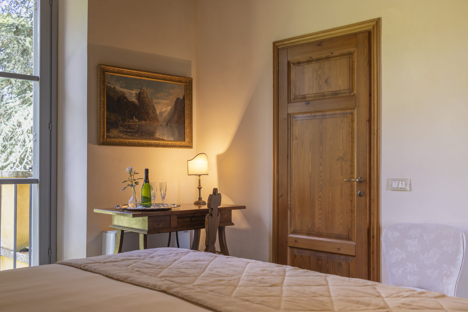 Cozy bedroom interior featuring a wooden door, a bedside table with a lamp and a bottle of wine, and a landscape painting on the wall, creating a warm and inviting atmosphere. | Only on SopranoVillas