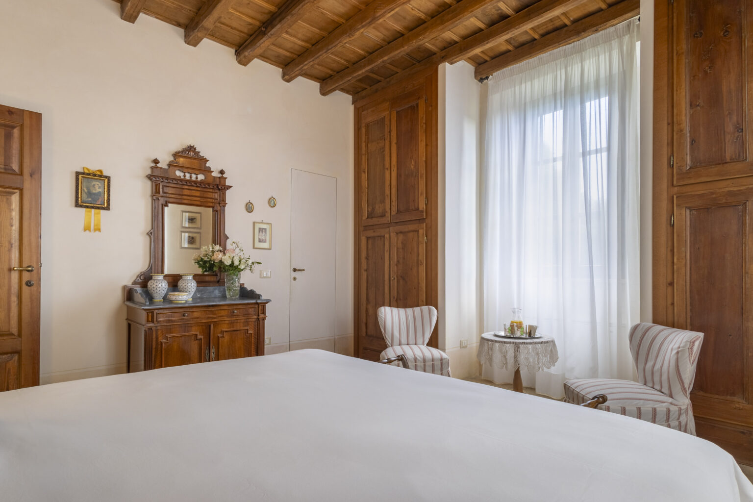 Cozy bedroom featuring wooden beams, elegant furnishings, and soft natural light filtering through sheer curtains. The room includes a comfortable bed, vintage-style armchairs, and a decorative mirror, creating a warm and inviting atmosphere. | Only on SopranoVillas