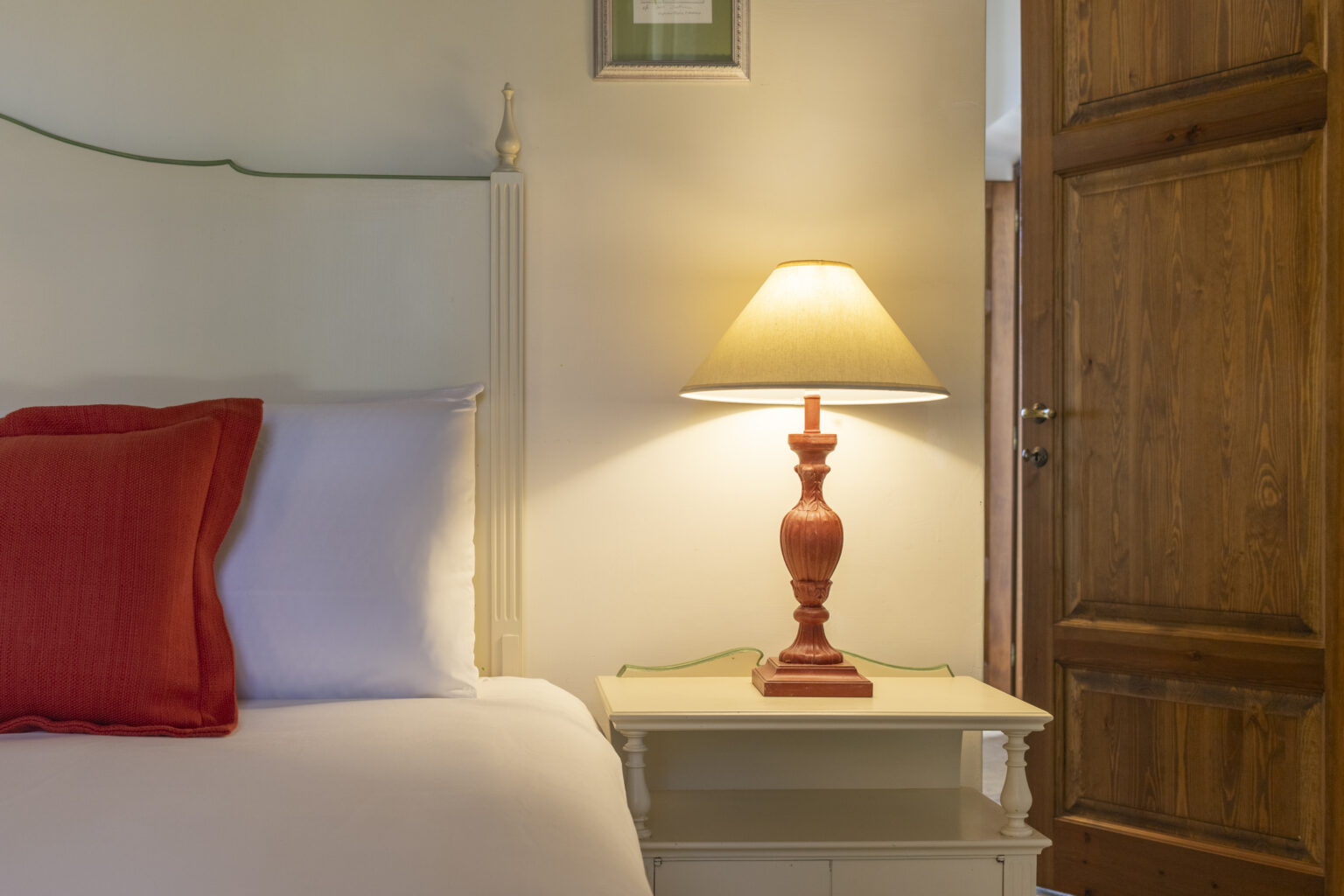 Cozy bedroom interior featuring a white bed with red accent pillows, a stylish nightstand with a red lamp, and a wooden door, creating a warm and inviting atmosphere. | Only on SopranoVillas