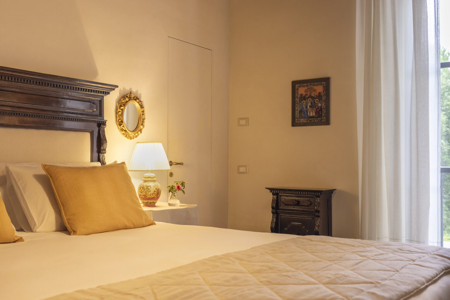 Cozy bedroom interior featuring a wooden bed frame with decorative pillows, a bedside lamp, and a small nightstand. Natural light filters through sheer curtains, highlighting warm tones and elegant decor. | Only on SopranoVillas