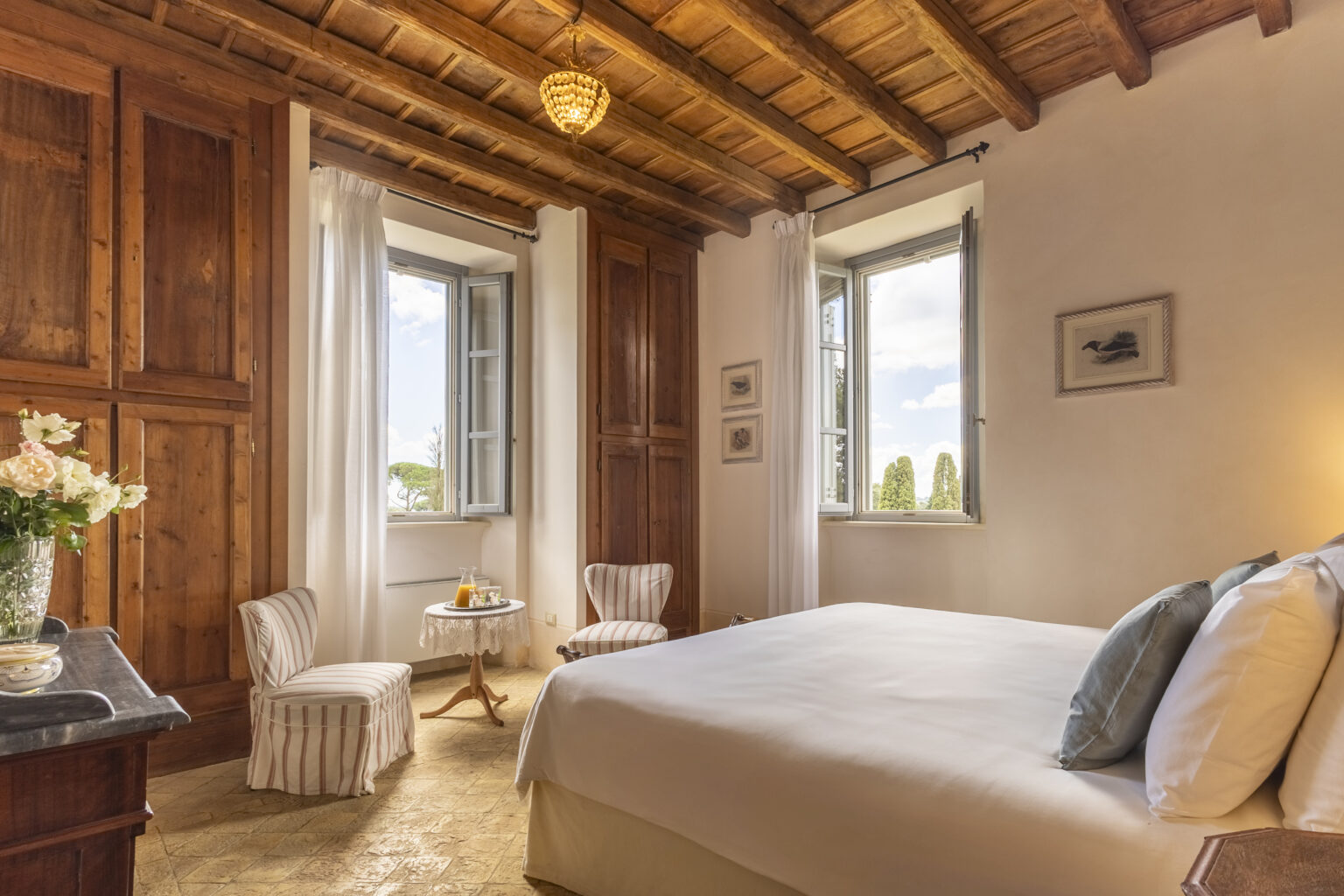 Cozy bedroom featuring a queen-sized bed, wooden ceiling beams, and large windows with scenic views. The room includes elegant decor with a nightstand, two armchairs, and a small table, creating a warm and inviting atmosphere. | Only on SopranoVillas