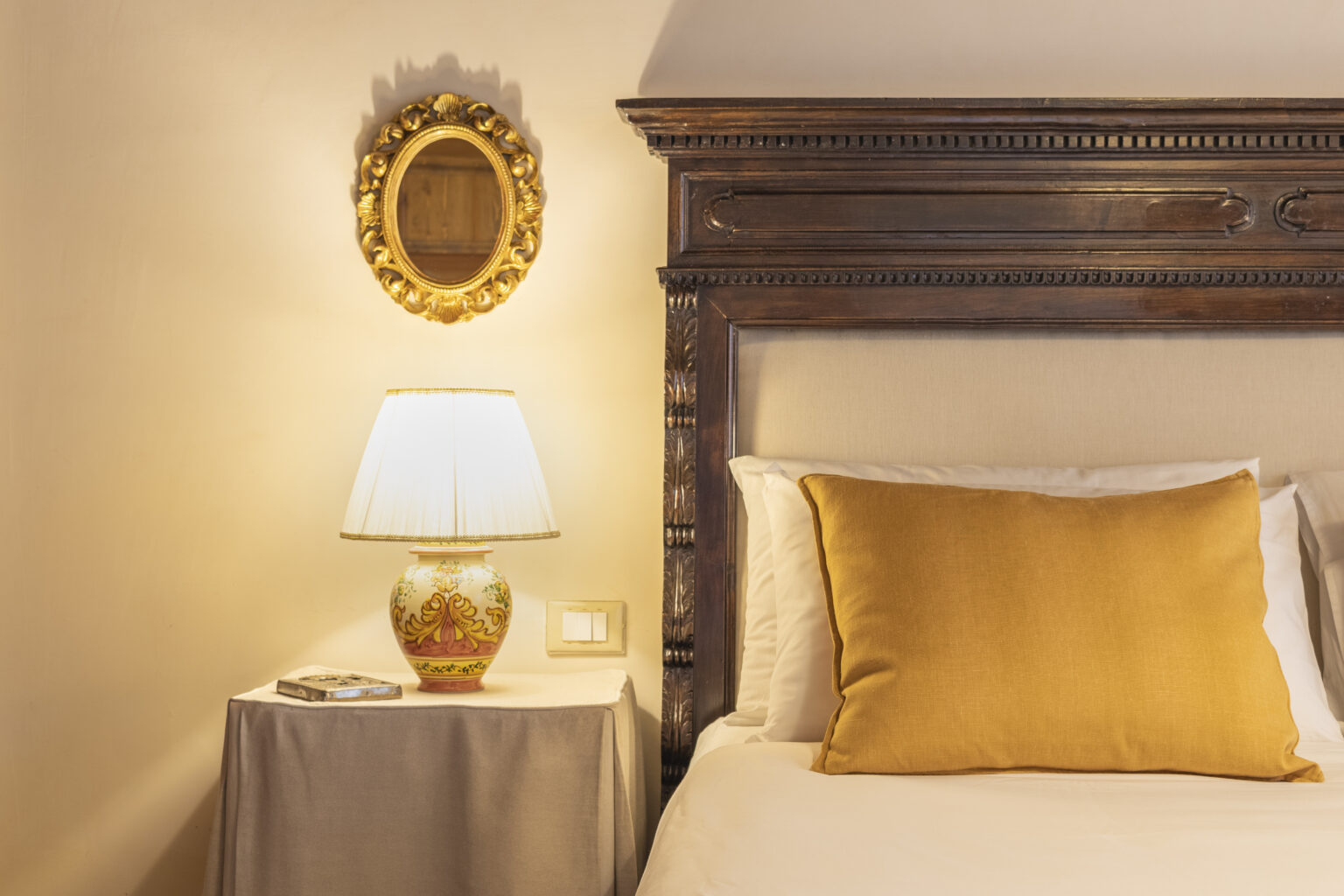 Cozy bedroom interior featuring a wooden bed frame, a gold-accented lamp on a nightstand, and a decorative mirror on the wall, creating a warm and inviting atmosphere. | Only on SopranoVillas