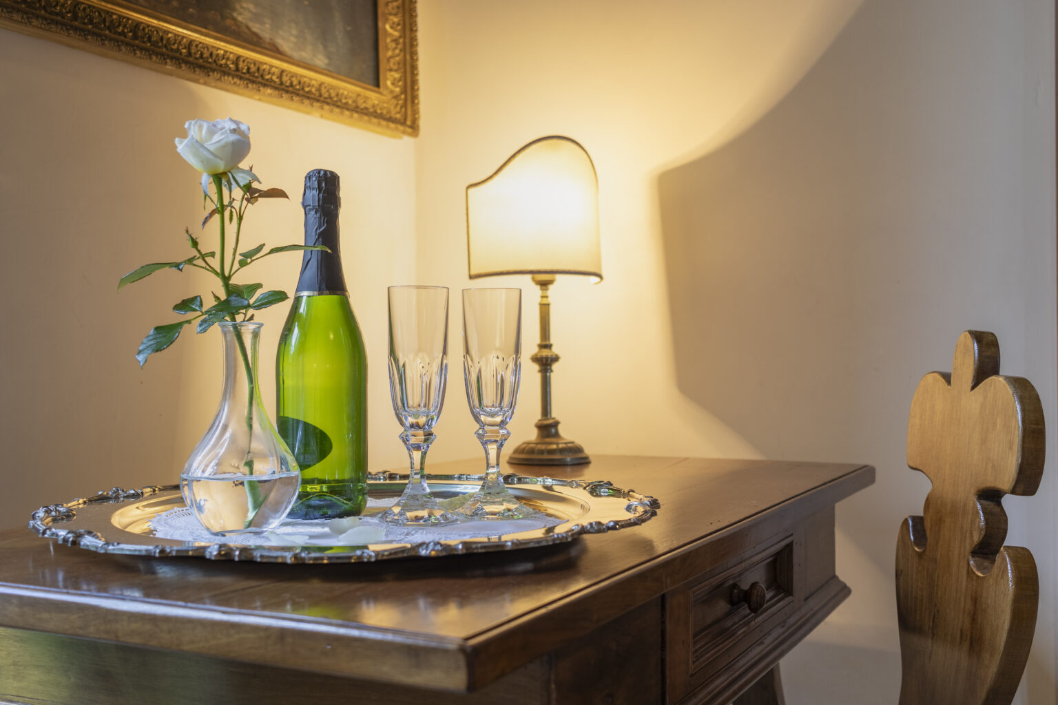 Elegant table setting featuring a green sparkling wine bottle, two crystal champagne glasses, and a single white rose in a vase on a silver tray, illuminated by a warm lamp in a cozy room. | Only on SopranoVillas