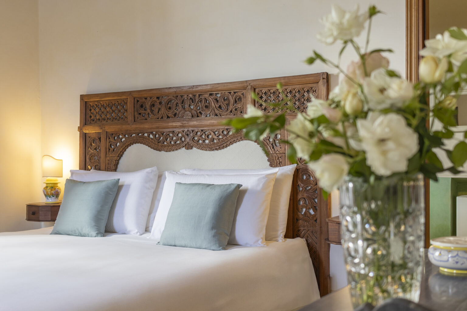 Elegant bedroom featuring a carved wooden headboard, white linens, and soft green accent pillows. A bedside lamp and a vase of fresh flowers add warmth and charm to the inviting space. | Only on SopranoVillas