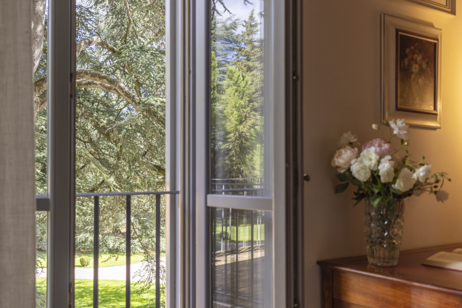 Interior view of a cozy room featuring a window with a balcony overlooking a lush green landscape, complemented by a vase of fresh flowers on a wooden dresser and a framed painting on the wall. | Only on SopranoVillas