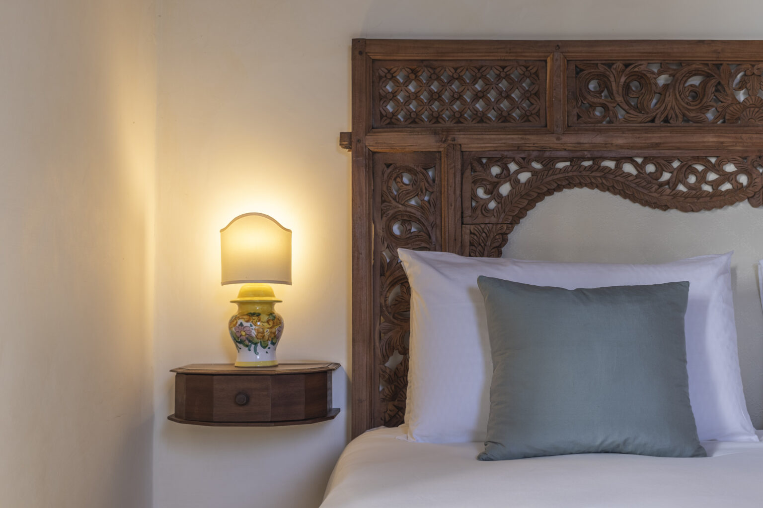 Cozy bedroom featuring a wooden headboard with intricate carvings, a white bed with a soft green pillow, and a bedside table with a decorative lamp, creating a warm and inviting atmosphere. | Only on SopranoVillas