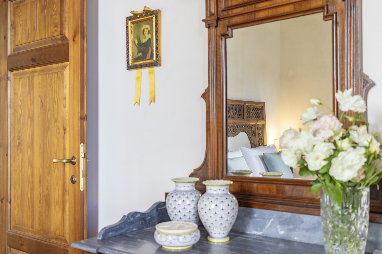 Elegant bedroom interior featuring a wooden door, ornate mirror, and decorative vases. A vintage portrait hangs on the wall, complemented by a floral arrangement in a glass vase. Soft lighting enhances the serene atmosphere, making it a perfect retreat. | Only on SopranoVillas
