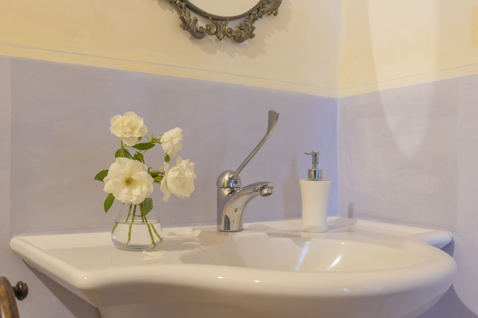 Elegant bathroom sink with a modern faucet, featuring a small vase of white roses and a soap dispenser, set against a light blue wall and adorned with a vintage mirror. | Only on SopranoVillas