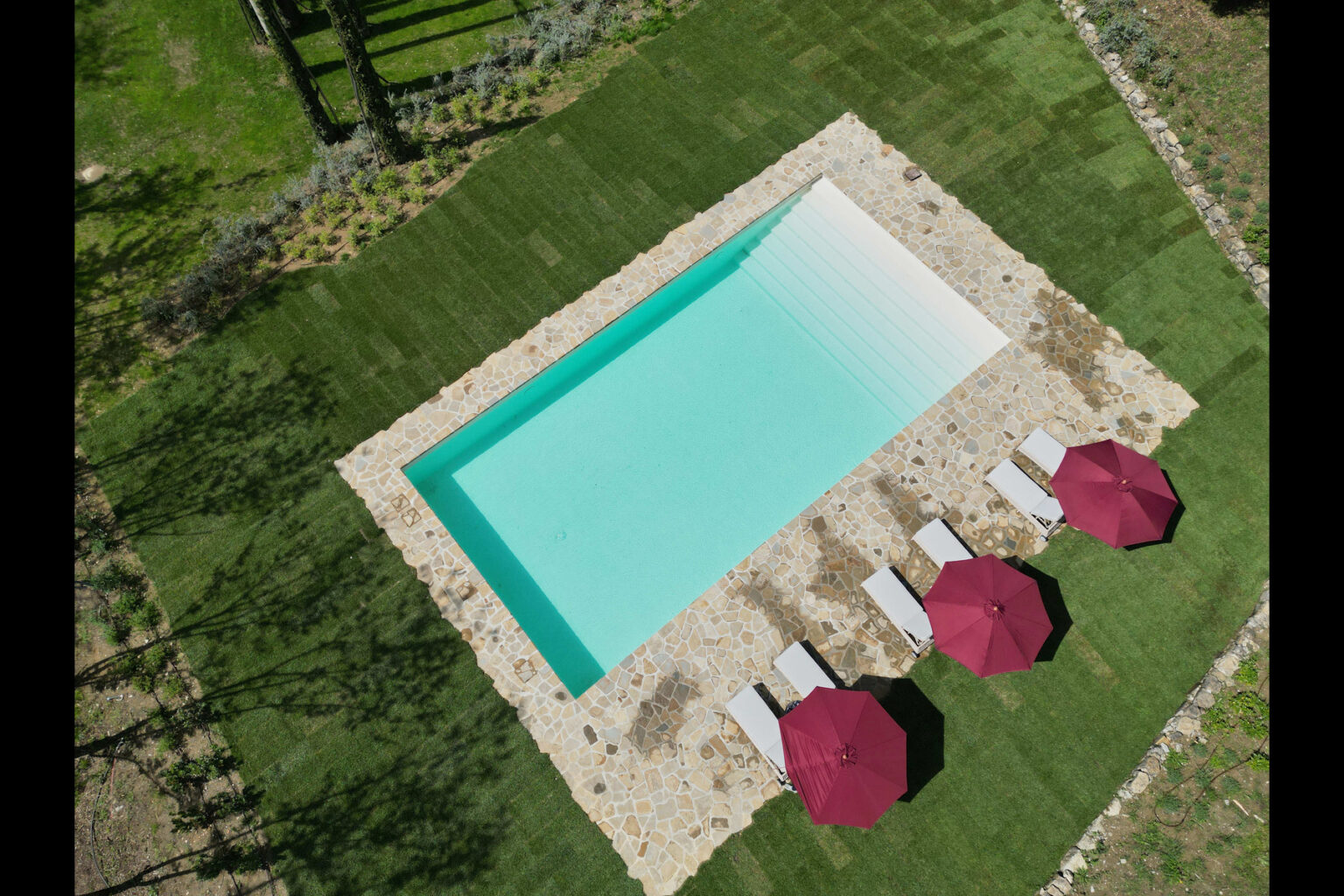 Aerial view of a modern swimming pool surrounded by lush green grass, featuring white lounge chairs and maroon umbrellas for shade. Ideal for outdoor relaxation and summer gatherings. | Only on SopranoVillas