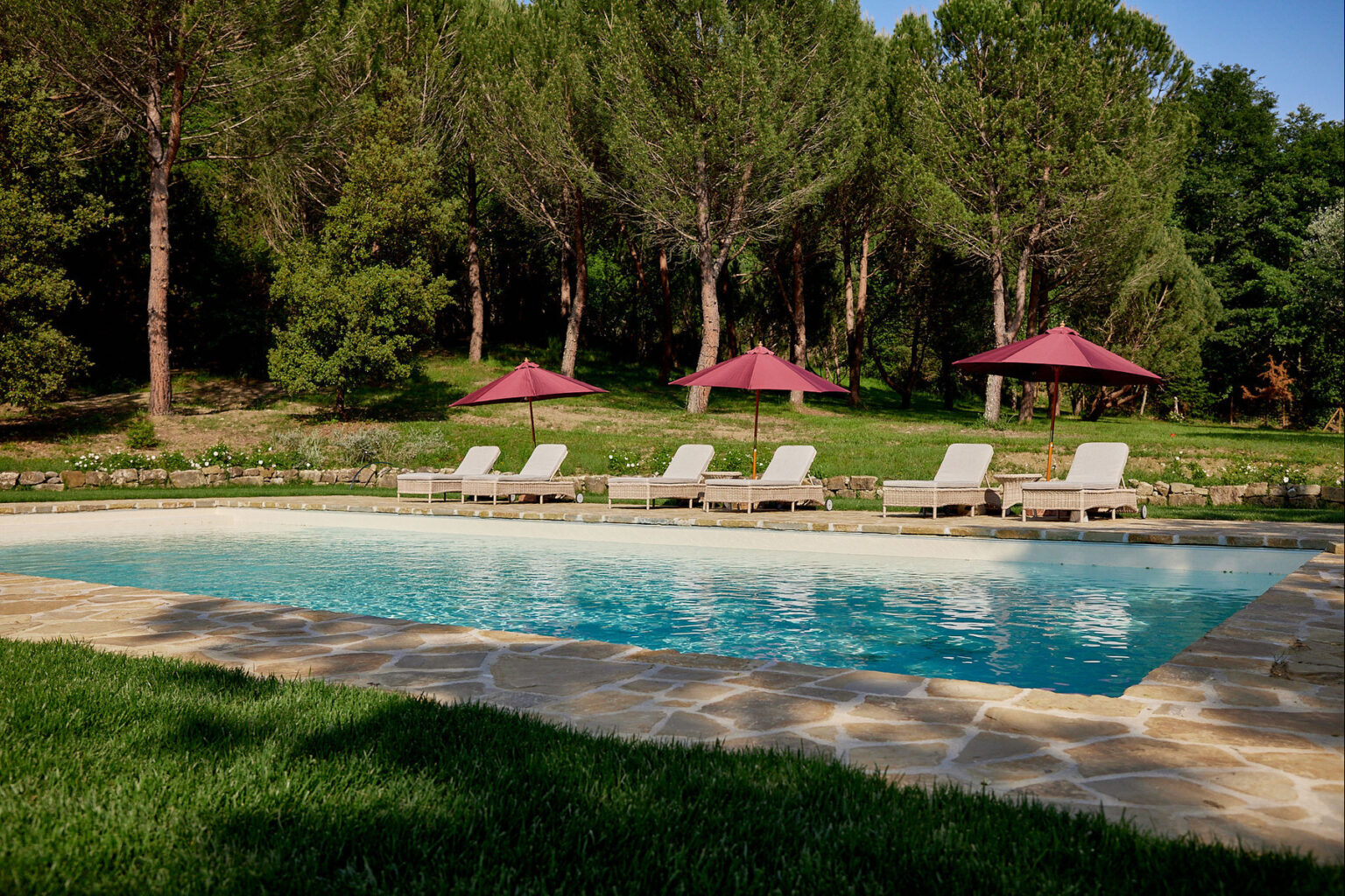 Serene outdoor swimming pool surrounded by lush greenery, featuring comfortable lounge chairs and shaded cabanas for relaxation. Ideal for a tranquil getaway or summer retreat. | Only on SopranoVillas