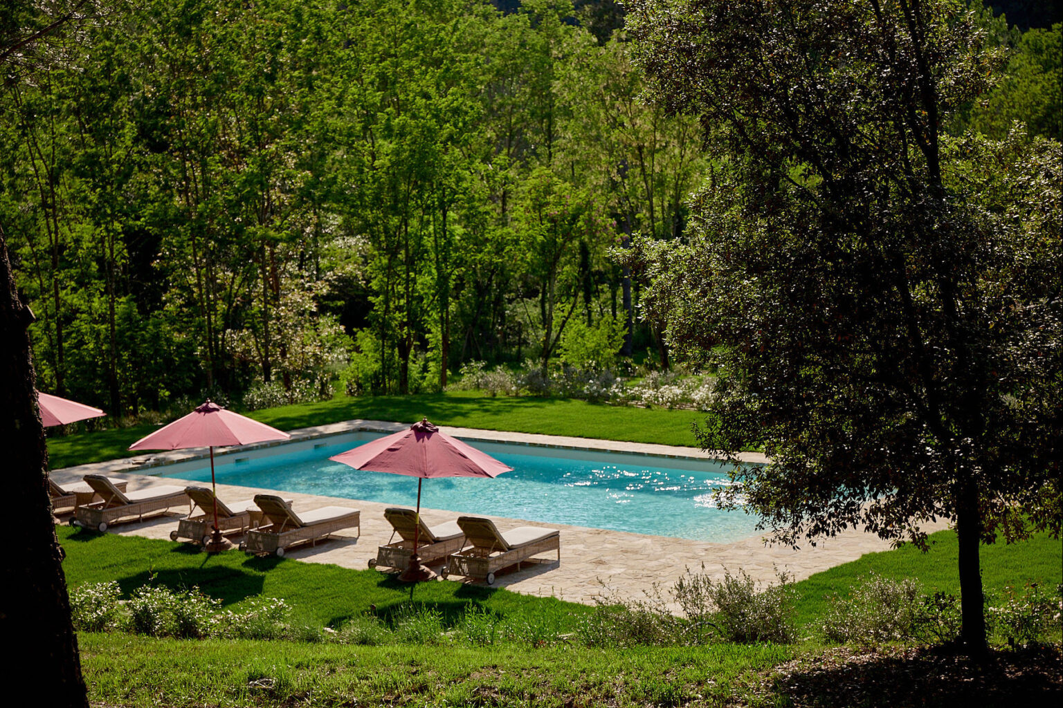 Serene outdoor swimming pool surrounded by lush greenery, featuring comfortable lounge chairs and pink umbrellas for shade, ideal for relaxation and leisure. | Only on SopranoVillas